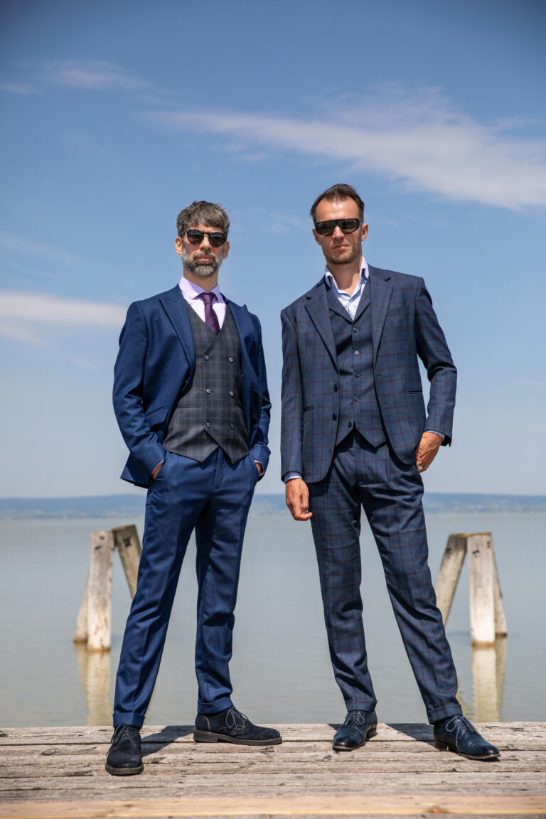 Zwei Männer in blauen Herrenanzügen mit Sonnenbrille stehen selbstbewusst auf einem Holzsteg am Meer